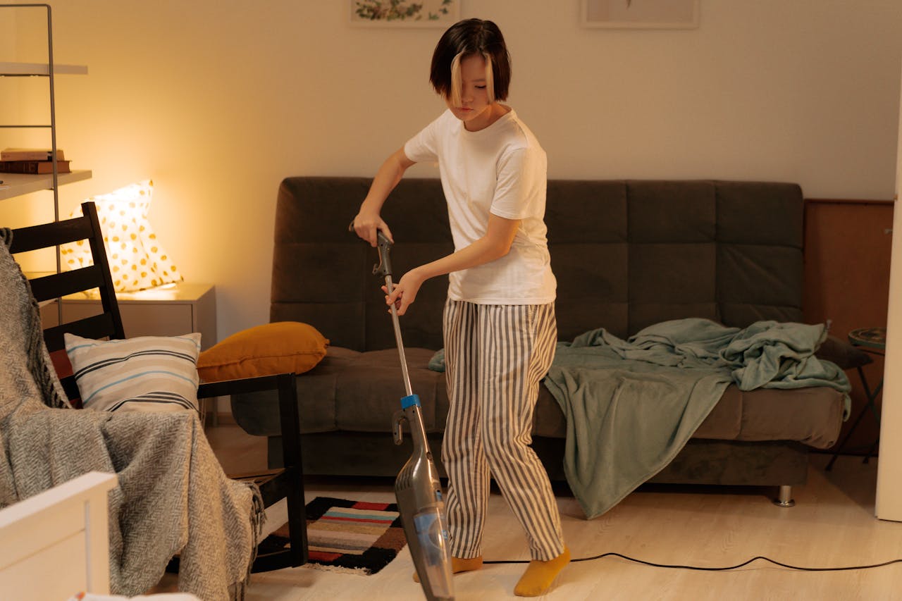 Asian woman cleaning a messy living room at home with a vacuum cleaner, creating a tidy space.